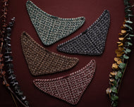 Four triangular knitted coasters in different colors on a dark brown background with leafy edges.