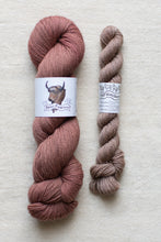 Load image into Gallery viewer, Two skeins of yarn, one pink and one brown, on a textured surface with labels.