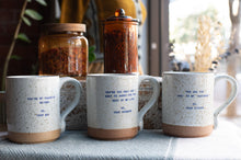Load image into Gallery viewer, Three speckled ceramic mugs with blue printed quotes are arranged on a tablecloth in front of two amber glass jars, with a softly blurred indoor background.