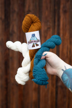 Load image into Gallery viewer, Three skeins of yarn in white, brown, and blue held by a hand against a wooden background.
