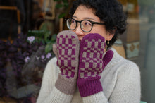 Load image into Gallery viewer, A person wearing a cream knit sweater and black-rimmed glasses holds up a pair of knitted mittens with a purple and beige checkerboard pattern in front of their face.
