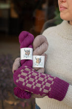 Load image into Gallery viewer, A person in a cream knit sweater holds two skeins of yarn and a knitted mitt. The mitt has a purple and beige checkerboard pattern with tassel ends.