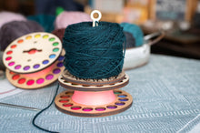 Load image into Gallery viewer, Spool of dark green thread on a wooden spool with colorful accents, placed on a textured fabric surface.