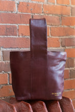 Load image into Gallery viewer, Brown leather tote bag with 'BE DESIGNED' branding against a brick wall.