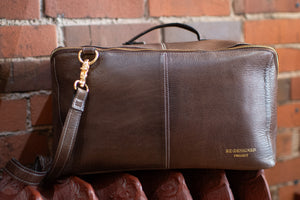 Brown leather bag with a gold zipper pull against a brick wall.