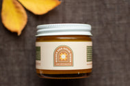 A jar of Golden Sun Serum with a white lid and cream label, set on a dark textured surface with yellow autumn leaves in the background.