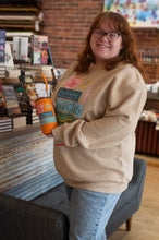 Load image into Gallery viewer, Person wearing a beige sweatshirt with text and graphics, holding a colorful cup in a casual setting.