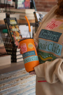 Person holding an orange tumbler with 'The Knitters Book Club' branding in a bookstore setting.