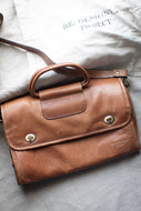 Brown leather bag on a light gray surface with 'RE-DESIGN PROJECT' text in the background.