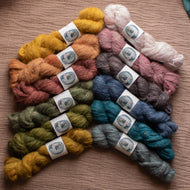 Six skeins of fluffy yarn arranged in a fan shape on a textured brown fabric background. The yarn colors include mustard yellow, peach, dusty rose, mauve, slate blue, teal, olive green, and heather gray.