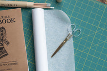 Load image into Gallery viewer, Scissors on a roll of paper with a notebook and pencils on a grid surface
