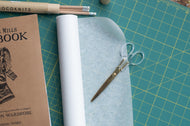 Scissors on a roll of paper with a notebook and pencils on a grid surface