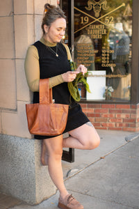 Woman holding a brown leather tote bag in front of a store window.
