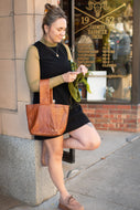 Woman holding a brown leather tote bag in front of a store window.