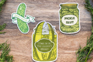Three pickle-themed stickers on a wooden surface with herbs.