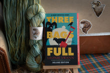 Load image into Gallery viewer, Book titled &#39;Three Bags Full&#39; by Leonie Swann next to skeins of yarn on a wooden surface.