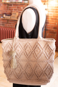 Beige textured tote bag with tassel on a mannequin against a brick wall background