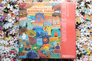 Jigsaw puzzle box with colorful design of national parks and scattered puzzle pieces on a wooden surface.