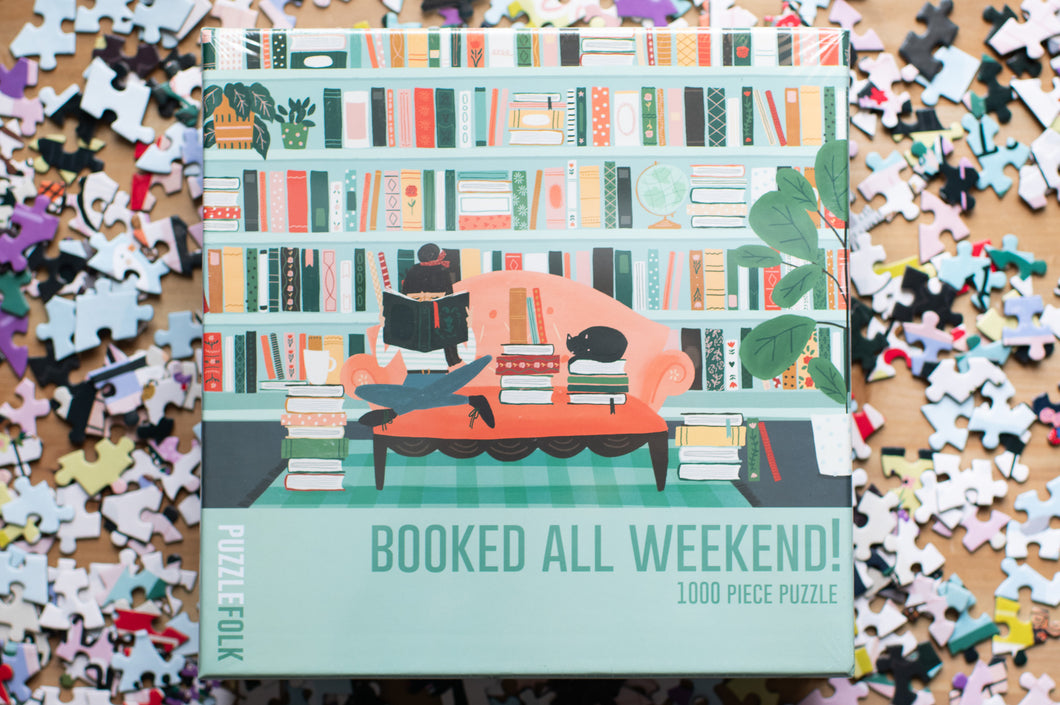 Jigsaw puzzle box with a design of people on a couch reading books, surrounded by scattered puzzle pieces.