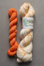 Load image into Gallery viewer, Two skeins of yarn, one orange and one multicolored with a label, on a gray background.