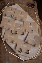Load image into Gallery viewer, Set of gold earrings on SONÓQ branded cards displayed on a wooden surface.