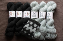 Load image into Gallery viewer, Three skeins of black yarn, one checkered sock, and two skeins of gray yarn on a wooden surface.