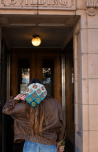 Load image into Gallery viewer, A person viewed from behind wearing a brown leather jacket and a colorful crocheted beanie with geometric patterns in blue, green, orange, and white, standing in front of a building entrance.