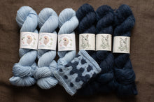 Load image into Gallery viewer, Set of blue and navy yarn skeins with matching knitted fabric on a brown background