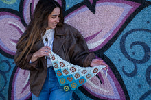 Load image into Gallery viewer, A woman wearing a brown leather jacket and blue jeans holds up a crocheted triangle shawl with a colorful geometric pattern, set against a vibrant graffiti wall.