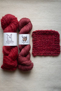 Two skeins of red yarn with labels and a knitted swatch on a beige surface