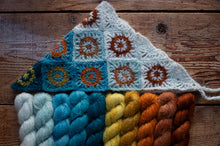 Load image into Gallery viewer, A handmade crocheted or knitted triangular textile piece with granny-square panels in white, blue, orange, and yellow, displayed on a rustic wooden surface above matching twisted yarn skeins in white, blue, yellow, and orange.