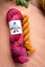 Load image into Gallery viewer, Two skeins of yarn, one pink and one orange, on a textured surface with a brand label visible.