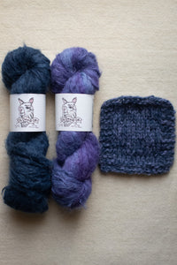 Two skeins of blue and purple yarn with labels and a knitted sample on a beige background