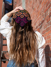 Load image into Gallery viewer, A woman viewed from behind wearing a crocheted beanie with a multicolor geometric pattern, standing against a red brick wall.