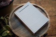White notebook titled 'The Knitwear Manual' on a wooden surface with plants in the background
