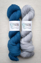 Load image into Gallery viewer, Two skeins of yarn, one blue and one gray, with labels on a beige background.