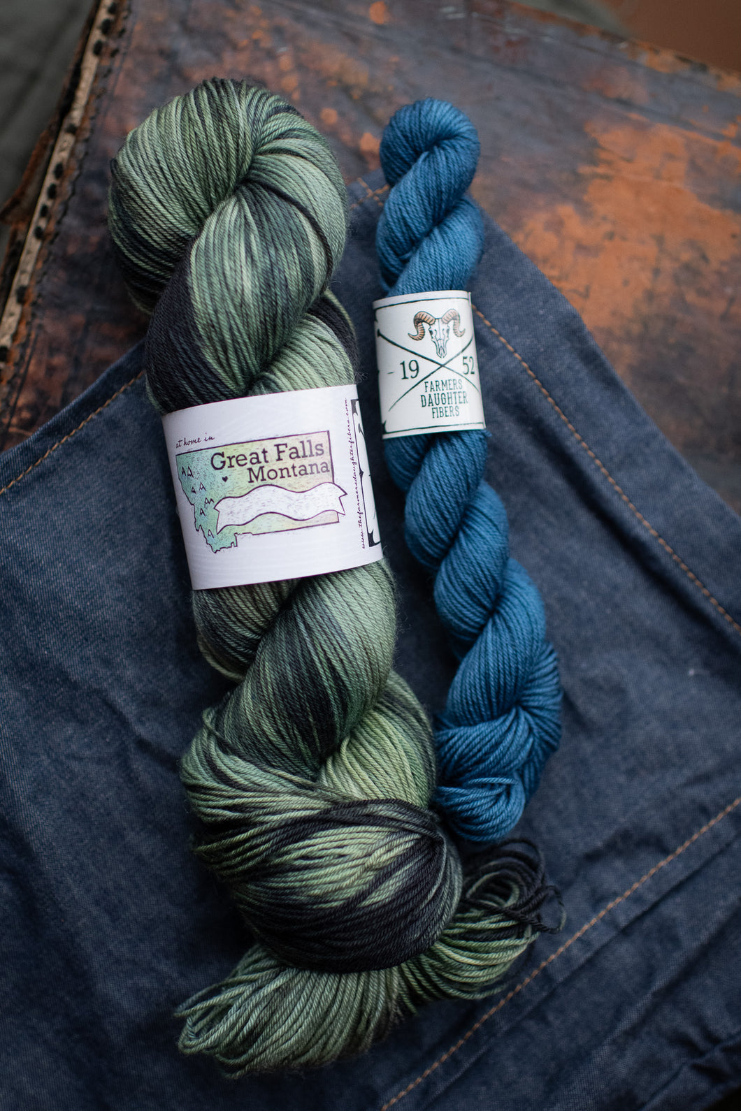 Two skeins of yarn, one green and one blue, on a denim surface.