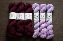 Load image into Gallery viewer, Two sets of yarn skeins, one in dark red and the other in purple with blue speckles, on a brown surface.