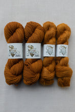 Load image into Gallery viewer, Four skeins of yarn in warm rust and camel shades, arranged side by side on a light gray textured fabric background. Each skein is wrapped with a white paper label featuring a ram illustration and the brand name 'Fishburn'.