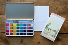 Load image into Gallery viewer, A watercolor paint palette with 36 color pans, an open lid, a brush, a blank sketch sheet, and a finished landscape watercolor painting, arranged on a rustic wooden tabletop.