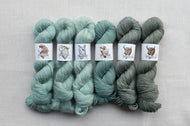 Six skeins of yarn in varying shades of green with labels on a gray background