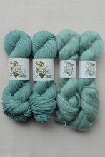 Load image into Gallery viewer, Four skeins of mint green yarn with white paper labels wrapped around them, arranged side by side on a light beige textured fabric background. The labels show the brand name 'Pimècus'.