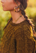 Load image into Gallery viewer, A close-up side profile of a woman wearing a textured olive-brown cable-knit sweater, gold hoop earrings, and a gold chain necklace against a warm, blurred outdoor background.