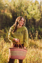 Load image into Gallery viewer, A woman standing in a grassy outdoor field wearing a textured olive green sweater and holding a pink and beige woven straw tote bag.