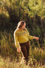 Load image into Gallery viewer, A woman standing in a sunlit grassy field, wearing a mustard yellow knit sweater with a dark pattern across the chest and brown pants.