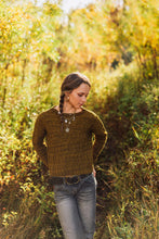 Load image into Gallery viewer, A woman wearing a textured olive-brown cable-knit sweater and gray jeans stands on a sunlit path surrounded by tall grass and autumn foliage.
