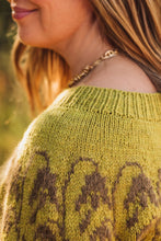 Load image into Gallery viewer, Close-up of a woman wearing a chunky knit sweater in olive green with a brown damask-style pattern, featuring a gold chain necklace against a warm, blurred outdoor background.
