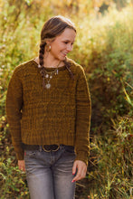 Load image into Gallery viewer, A woman wearing a textured olive-brown cable-knit sweater and gray jeans, standing outdoors in a sunlit grassy field.