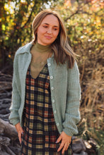 Load image into Gallery viewer, A woman wearing a sage green knit button-up cardigan over a brown and green plaid dress, standing outdoors in a wooded setting with autumn foliage.