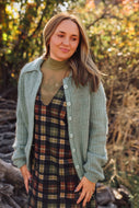 A woman wearing a sage green knit button-up cardigan over a brown and green plaid dress, standing outdoors in a wooded setting with autumn foliage.